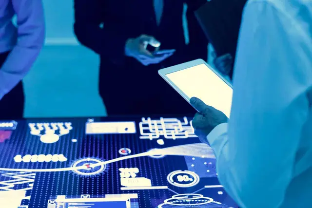 Pessoas com um tablet e um celular diante de uma mesa luminosa com gráficos e indicadores em tons de azul
