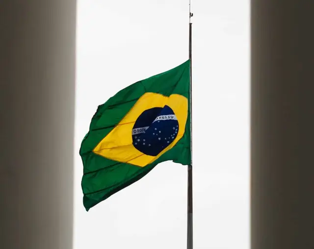 Bandeira do Brasil hasteada a meio mastro contra um céu branco, entre duas paredes escuras
