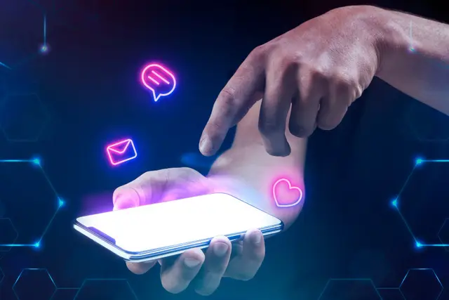 Mão tocando a tela de um celular com ícones de mensagem, e-mail e coração em neon rosa flutuando sobre fundo azul escuro.