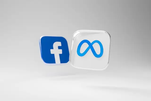 Dois ícones 3D flutuando sobre fundo cinza claro: o logotipo azul do Facebook e o símbolo de infinito da Meta em um cubo translúcido.