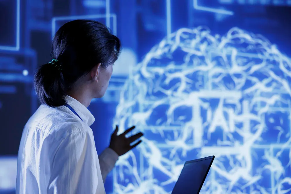 Mulher de perfil que levanta uma mão diante de um grande cérebro luminoso em azul, com um notebook em primeiro plano e a sigla AI na imagem.