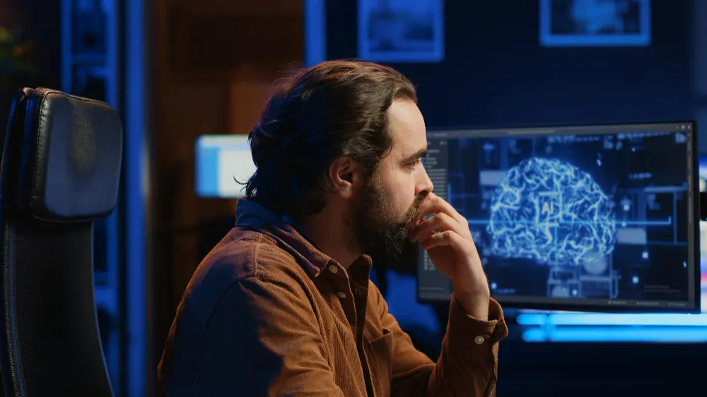 Homem barbudo com a mão na boca olha um monitor com a imagem de um cérebro azul, em um escritório de luz tênue.