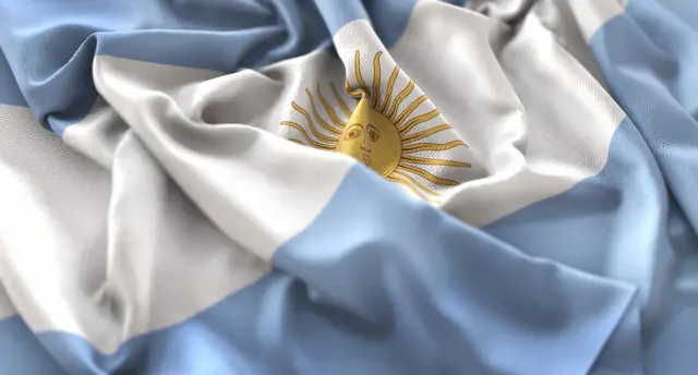 Close da bandeira argentina amassada, com faixas azul-claras e brancas e o Sol de Maio ao centro.