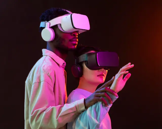 Duas pessoas com óculos de realidade virtual e fones de ouvido sob luz rosa e azul; uma levanta a mão como se tocasse algo à frente.