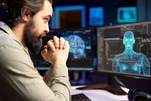 Homem barbudo de perfil com as mãos entrelaçadas diante de dois monitores com um cérebro digital e uma figura humana em 3D