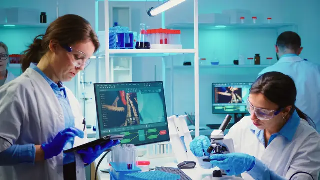 Duas cientistas de jaleco e luvas trabalham em um laboratório iluminado de azul, com um tablet, um microscópio e um monitor