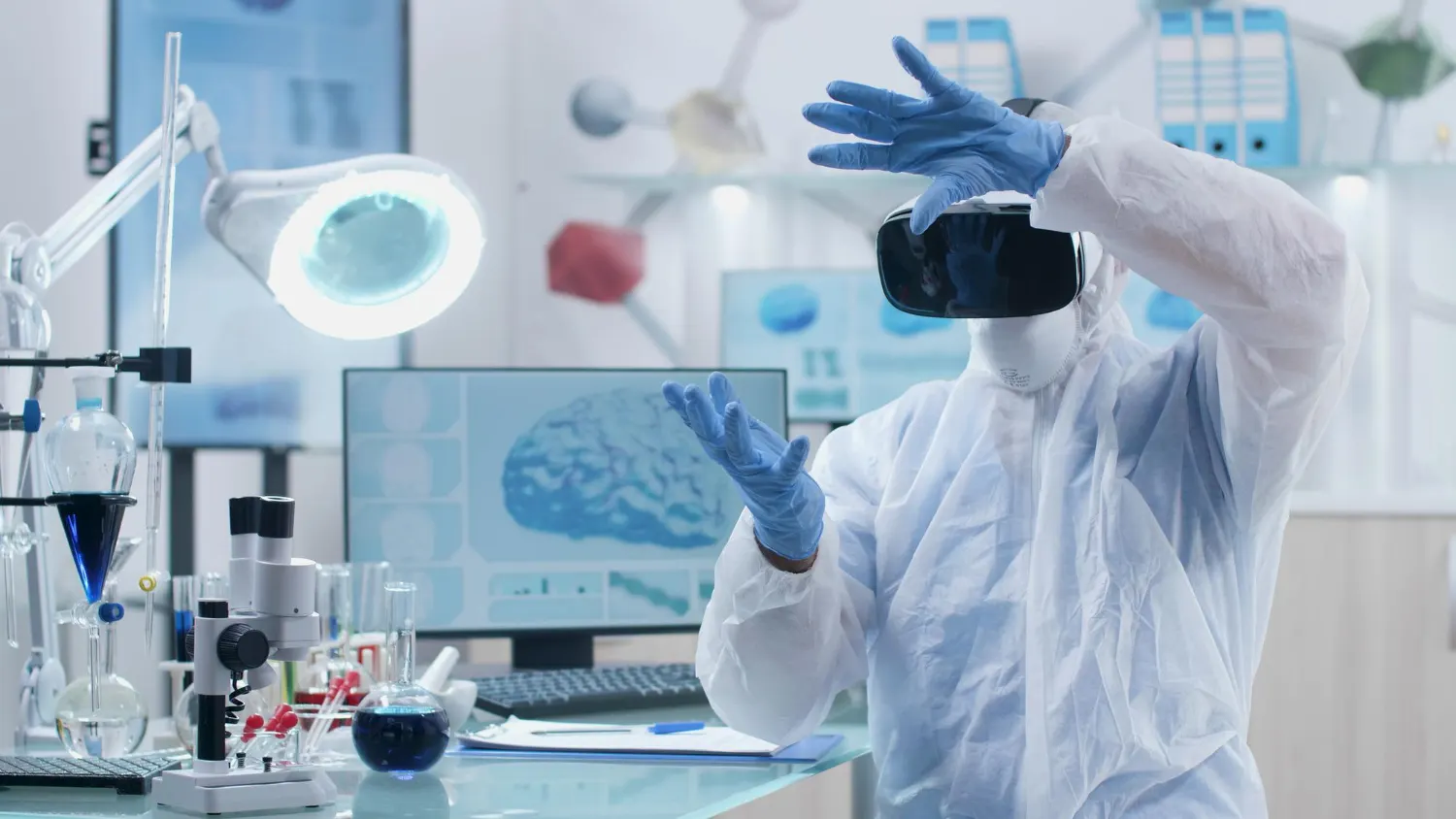 Pessoa com roupa de proteção e óculos de realidade virtual levanta as mãos enluvadas diante de um monitor que mostra um cérebro