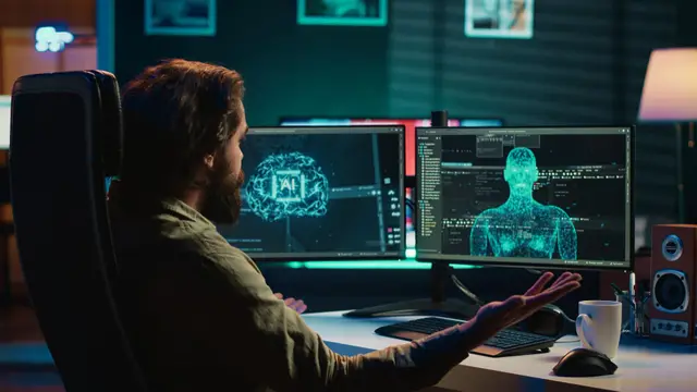 Homem barbudo diante de uma mesa, com as mãos abertas em frente a dois monitores com um cérebro digital e uma figura humana em 3D