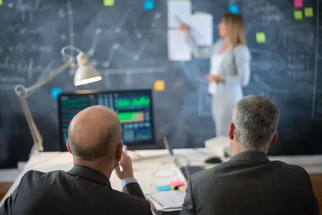 Dois homens de costas observam uma mulher que apresenta diante de um quadro-negro com anotações, com um monitor de gráficos em primeiro plano