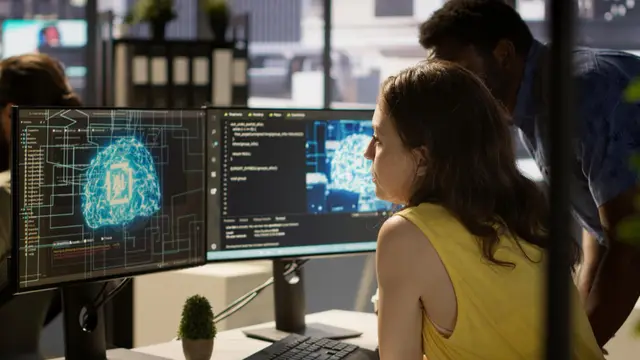 Mulher de blusa amarela e um homem atrás dela olham dois monitores com cérebros digitais e código em um escritório