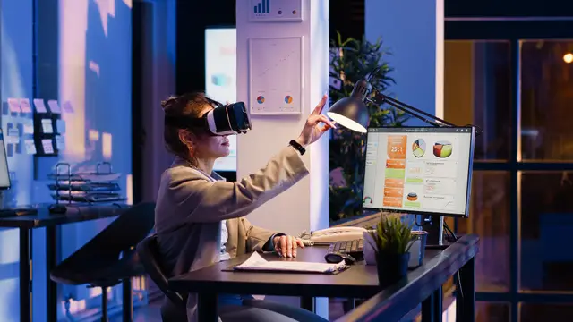 Mulher com óculos de realidade virtual levanta a mão diante de uma mesa com um monitor de gráficos, em um escritório à noite