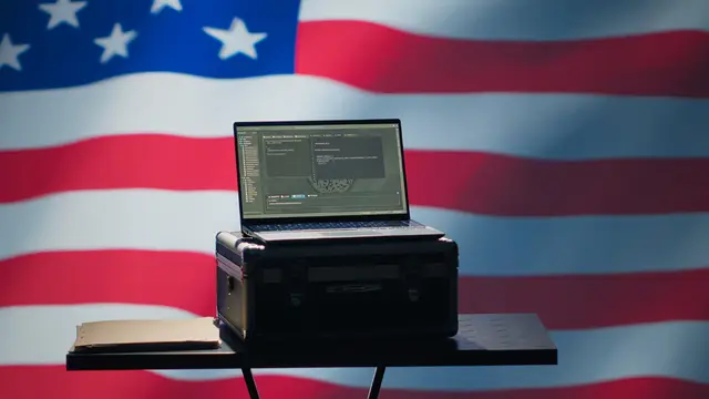 Laptop aberto sobre uma maleta preta em uma mesa pequena, diante de uma grande bandeira dos Estados Unidos.