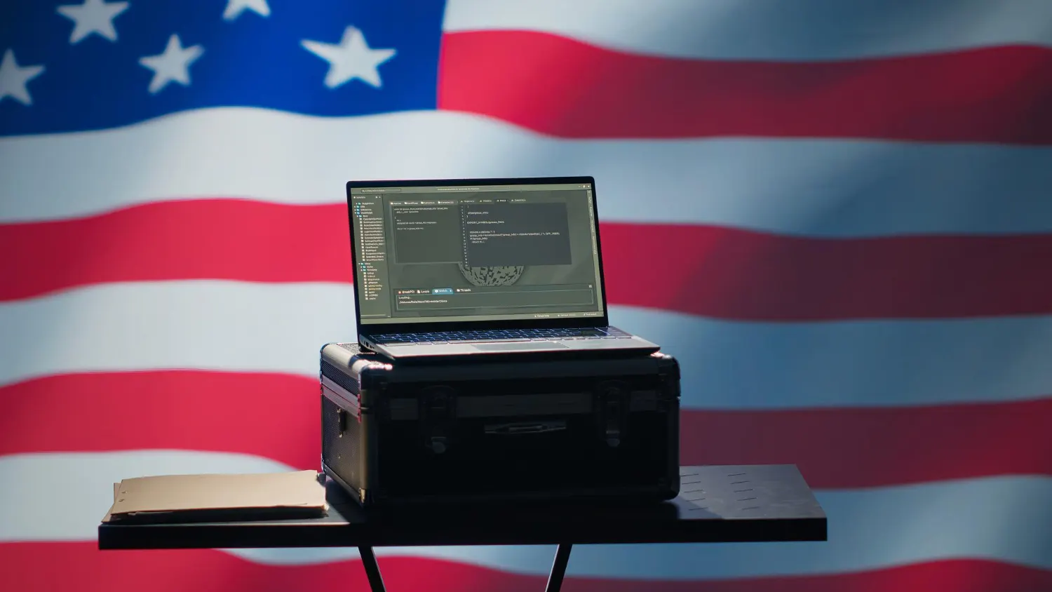 Laptop aberto sobre uma maleta preta em uma mesa pequena, diante de uma grande bandeira dos Estados Unidos.
