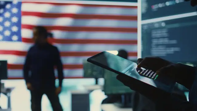 Silhueta de um homem diante de uma bandeira dos Estados Unidos e telas com código, com um tablet em primeiro plano