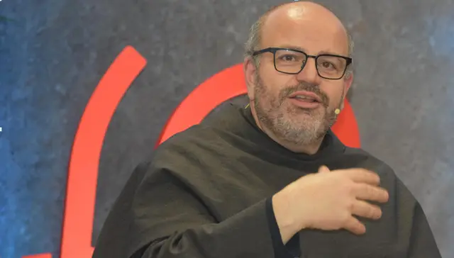 Close de um frade franciscano de óculos e hábito escuro que fala e gesticula diante de um fundo com formas vermelhas