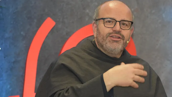 Close de um frade franciscano de óculos e hábito escuro que fala e gesticula diante de um fundo com formas vermelhas