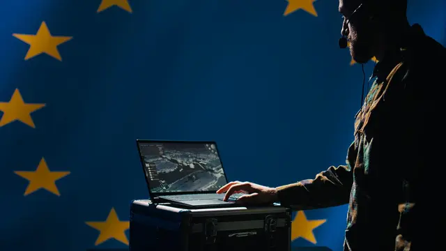 Homem de fones de ouvido digita em um laptop diante de uma bandeira azul da União Europeia com estrelas amarelas