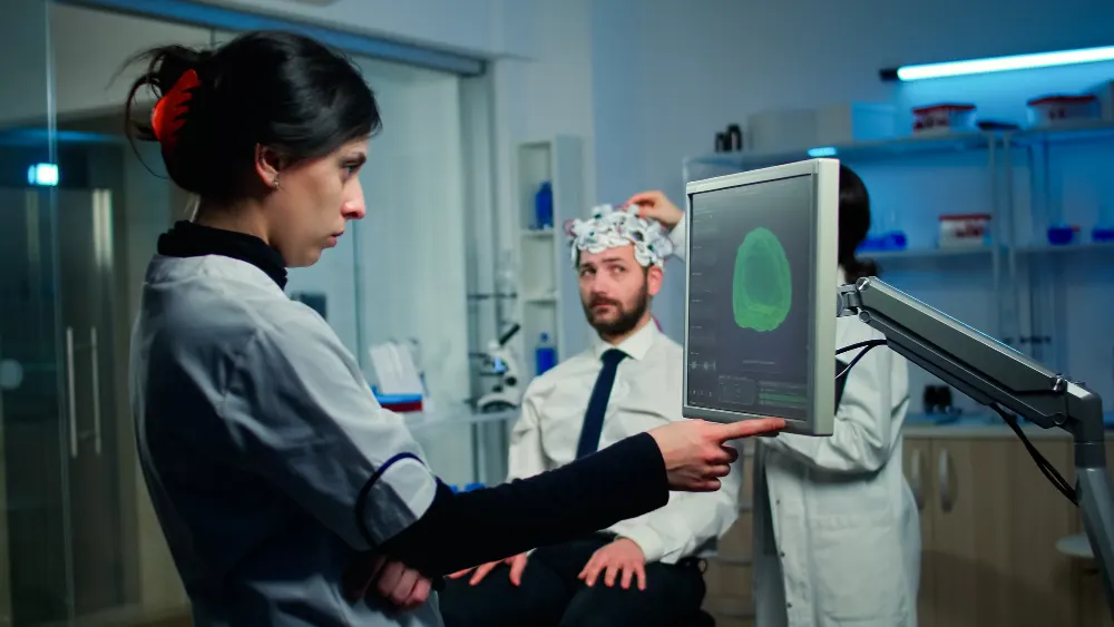 Pesquisadores em um laboratório apontam para uma tela com a imagem de um cérebro enquanto um colega usa uma touca de eletrodos
