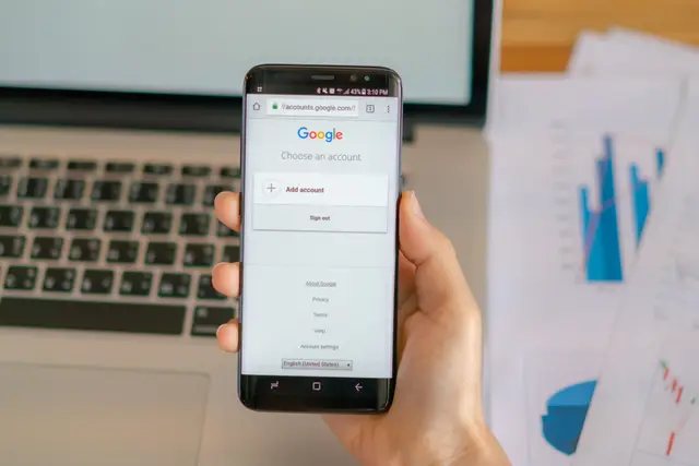 Mão segurando um celular com a tela de login do Google, ao lado de um notebook e gráficos impressos