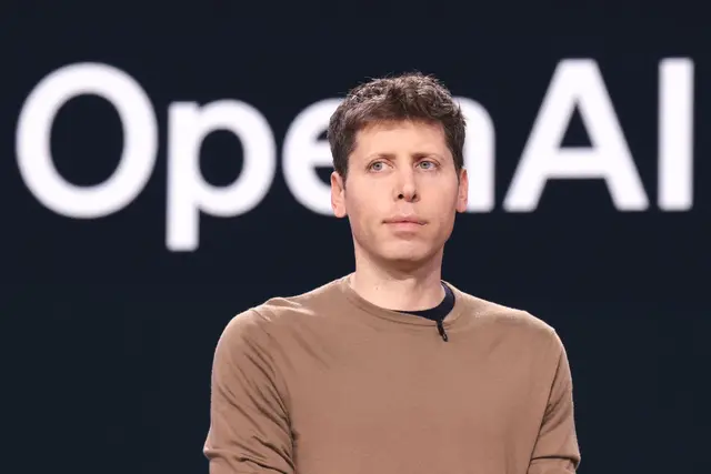 Sam Altman com camiseta marrom diante de um fundo escuro com o logotipo da OpenAI