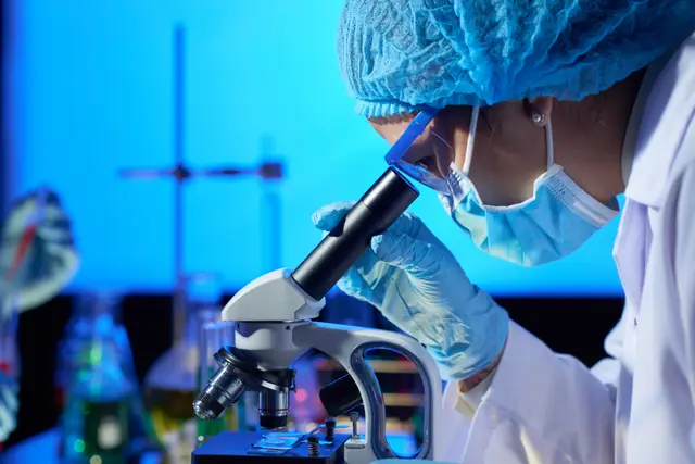 Pesquisadora com touca azul e máscara olha por um microscópio em um laboratório iluminado de azul