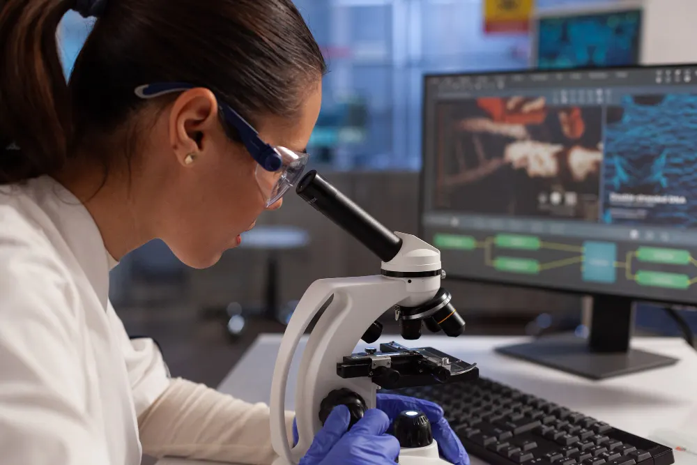 Pesquisadora de óculos de proteção olha por um microscópio, ao lado de um monitor com imagens de células, em um laboratório