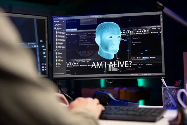 Monitor com uma cabeça digital azul e o texto «Am I alive?» diante de uma pessoa digitando, com mais telas de código ao redor