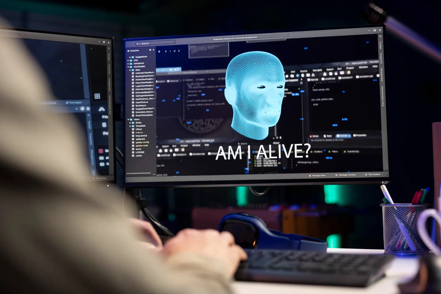 Monitor com uma cabeça digital azul e o texto «Am I alive?» diante de uma pessoa digitando, com mais telas de código ao redor