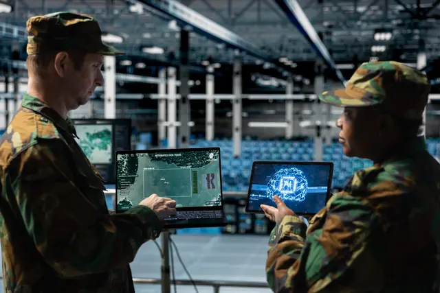 Dois militares de uniforme camuflado consultam mapas e uma tela com um cérebro digital em notebooks, dentro de um grande hangar