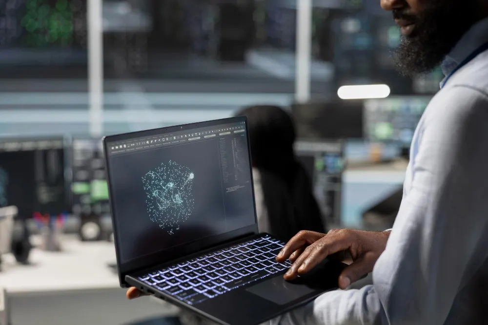 Homem de barba segura um notebook aberto que mostra uma rede de pontos luminosos, em uma sala com telas ao fundo