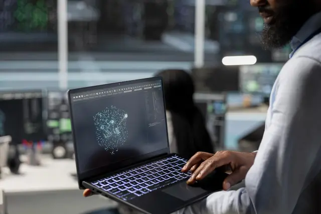 Homem de barba segura um notebook aberto que mostra uma rede de pontos luminosos, em uma sala com telas ao fundo