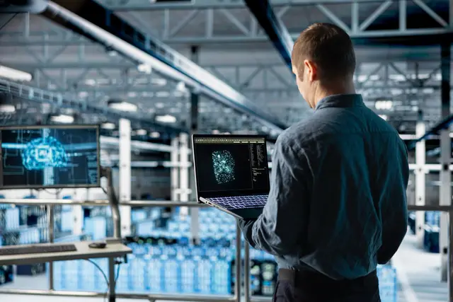 Homem visto de costas com um notebook aberto em um galpão industrial, ao lado de uma tela com uma figura digital azul