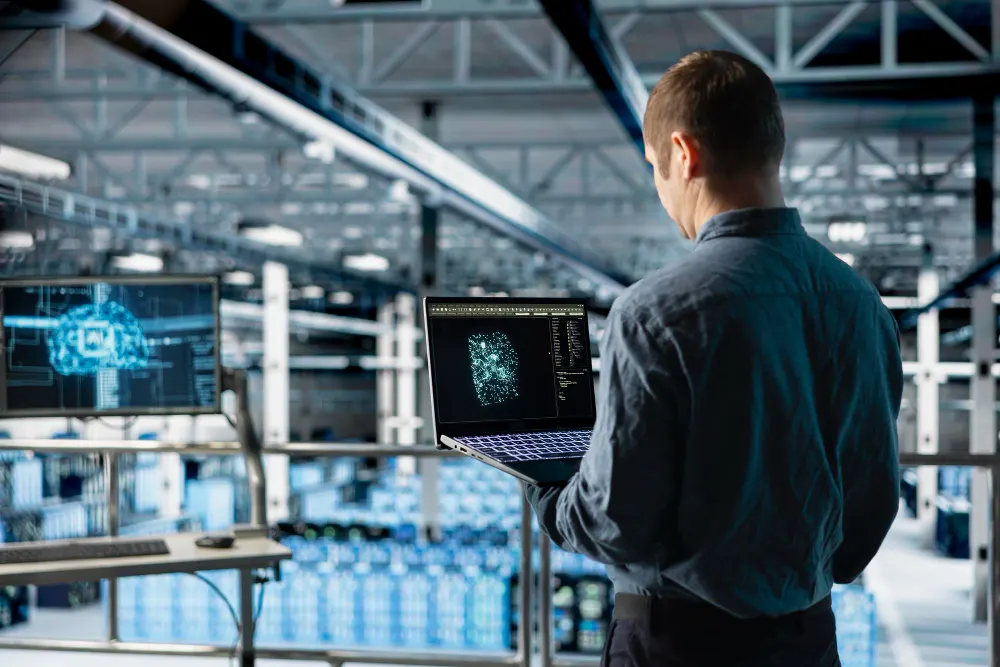 Homem visto de costas com um notebook aberto em um galpão industrial, ao lado de uma tela com uma figura digital azul