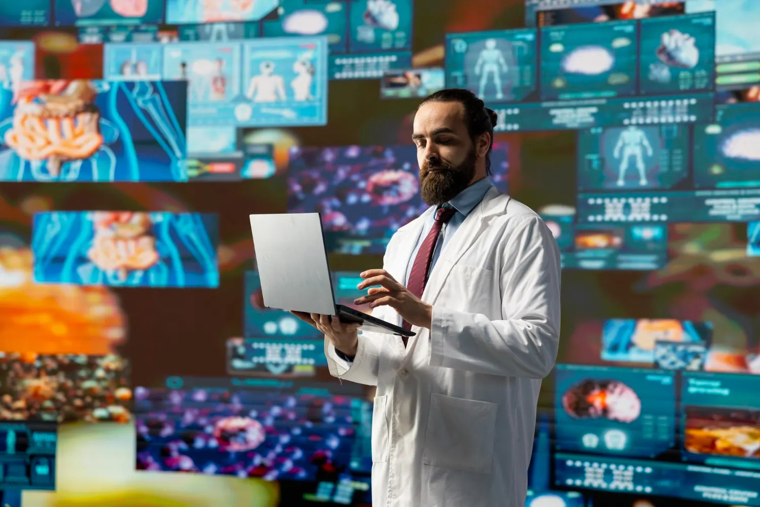 Médico de jaleco branco e gravata consulta um notebook diante de um painel de telas com imagens anatômicas e de células