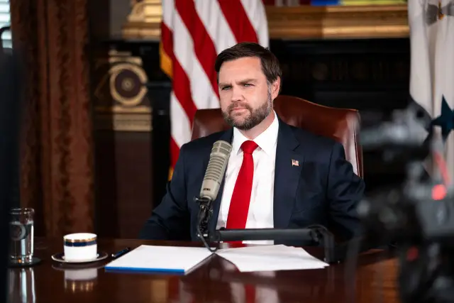 Homem de barba, terno escuro e gravata vermelha sentado diante de um microfone em um gabinete, com bandeiras americanas ao fundo