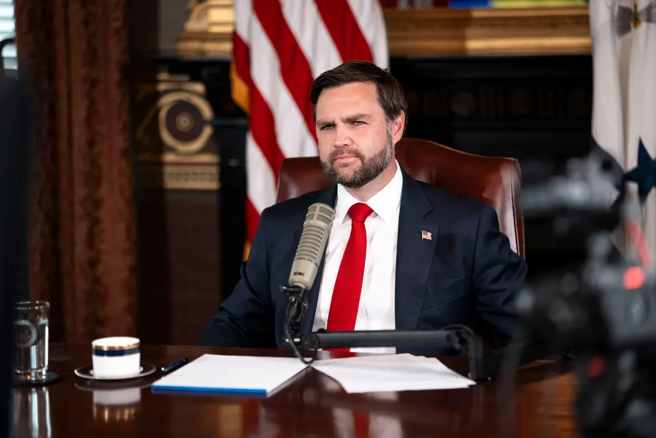 Homem de barba, terno escuro e gravata vermelha sentado diante de um microfone em um gabinete, com bandeiras americanas ao fundo