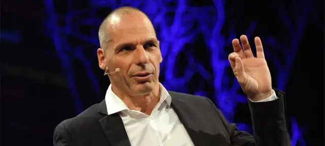 Yanis Varoufakis fala em um palco, com microfone de cabeça e uma das mãos erguida, diante de um fundo azul-escuro com luzes