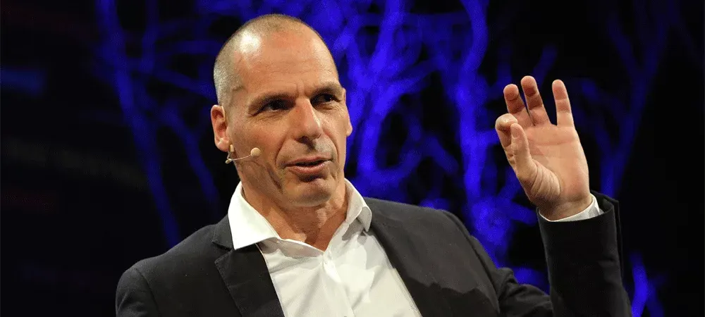 Yanis Varoufakis fala em um palco, com microfone de cabeça e uma das mãos erguida, diante de um fundo azul-escuro com luzes