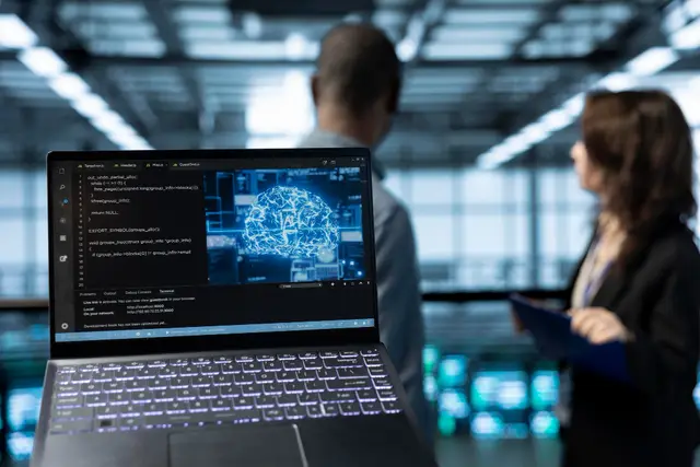 Notebook aberto com código e a imagem de um cérebro digital, com duas pessoas conversando desfocadas ao fundo de um escritório