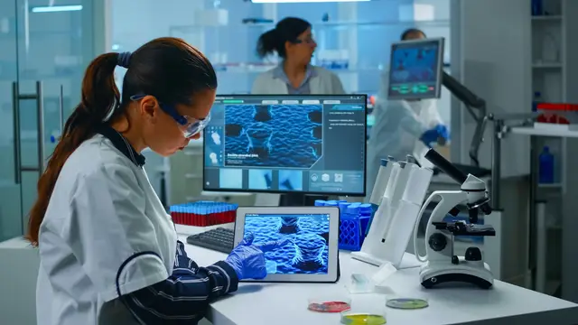 Cientista de jaleco, luvas e óculos de proteção olha para um tablet e um monitor com imagens microscópicas azuis em um laboratório
