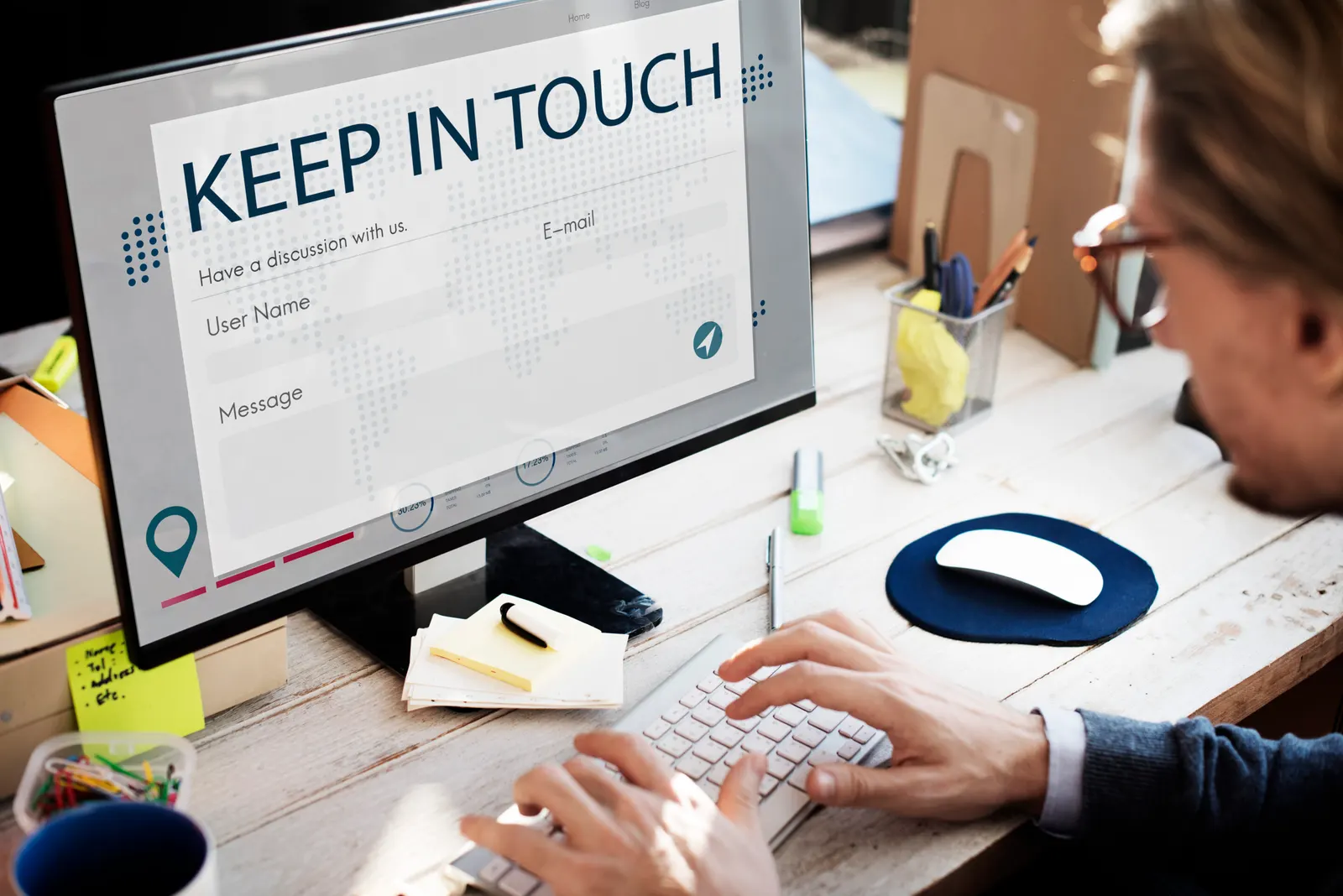 Homem de óculos digitando em um teclado diante de um monitor com um formulário de contato intitulado «Keep in touch»