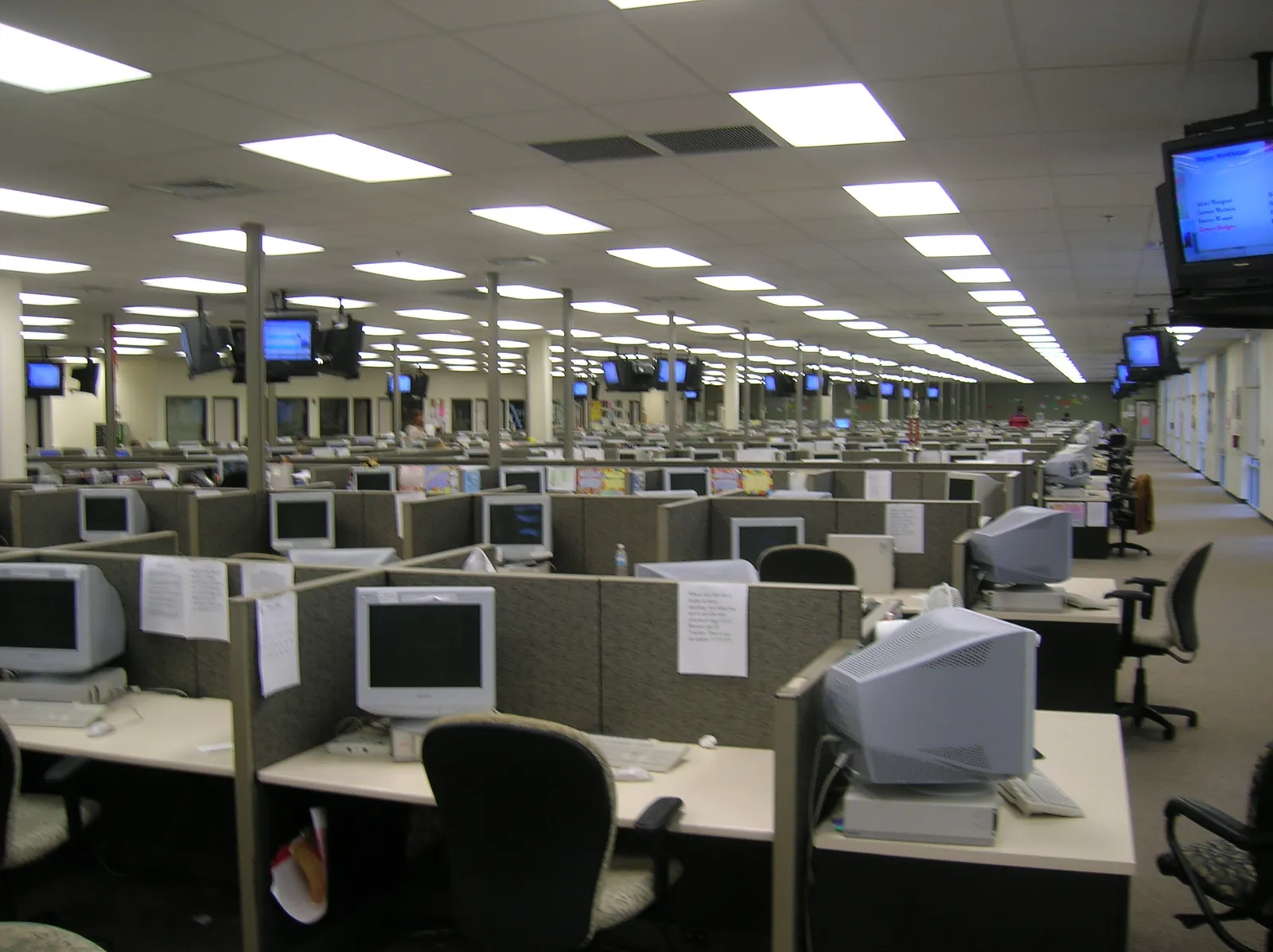 Fileiras de cubículos com monitores de tubo, teclados e cadeiras em um amplo escritório com luminárias retangulares no teto.